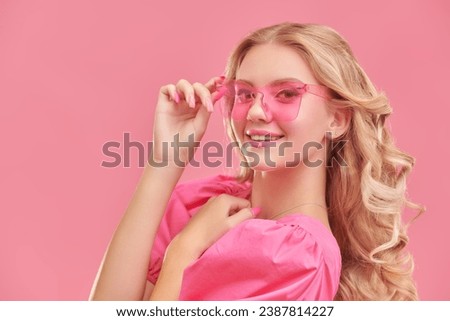 Femininity, beauty and fashion. A cute attractive girl with blond hair poses in a pink dress and pink glasses and smiles. Pink studio background with copy space. Young beauty. 