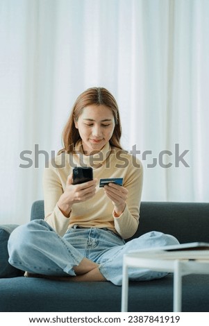 Cheerful Asian woman using smartphone, shopping online, holding credit card, paying, sitting on sofa at home Internet banking and e-commerce applications vertical picture