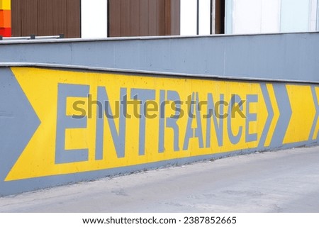 Entrance yellow sign with direction arrows printed on the gray concrete wall, horizontal photo.