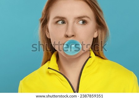 Beautiful young woman blowing bubble gum on light blue background Royalty-Free Stock Photo #2387859351