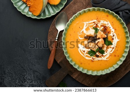 Pumpkin and carrot cream soup with herbs, seasonings and seeds in bowl on black wooden background in rustic style. Thanksgiving traditional autumn pumpkin cream soup. Top view. 