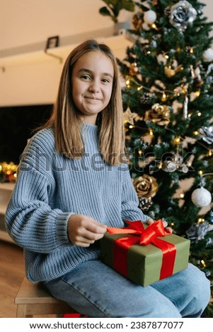 Vertical portrait of cute little girl holding wrapped with red ribbon and bow festive Christmas present gift box, sitting by glowing xmas tree in morning, smiling looking at camera.