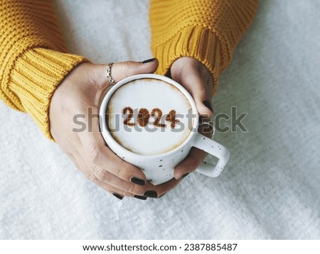 Happy New Year 2024 theme number 2024 over frothy surface of cappuccino served in white coffee mug holding by female hands on bed with white blanket background. Holidays food art, new year new you.