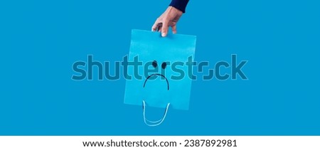 a man holding a blue shopping paper bag, with a sad face painted in it, upside-down on a blue background, in a panoramic format to use as web banner Royalty-Free Stock Photo #2387892981