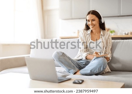 Young Smiling Woman In Headset Study Online With Laptop From Home, Happy Millennial Female Watching Webinar On Computer And Taking Notes To Notepad, While Sitting On Couch In Living Room Royalty-Free Stock Photo #2387959497