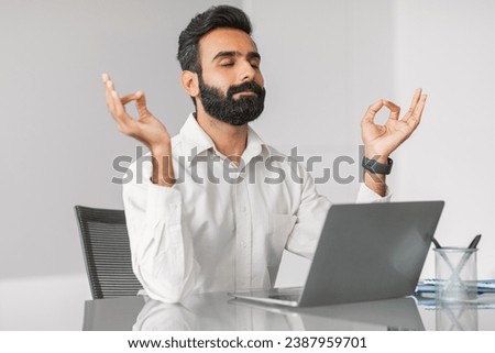 Calm indian businessman in white shirt practicing meditation at his office desk to alleviate stress, with eyes closed and fingers in mudra position, amidst busy workday Royalty-Free Stock Photo #2387959701