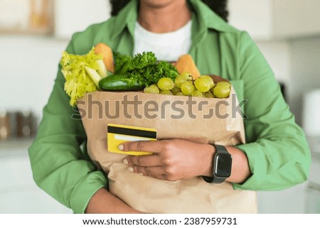 Cropped shot of African American woman holding her credit card and delivered grocery order in paper shopping bag, posing in modern kitchen interior. Unrecognizable buyer with food products