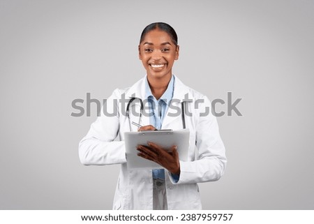 Cheerful millennial black lady doctor in white coat, with stethoscope, write recipe at clipboard, isolated on gray studio background. Medical exam, health care consultation, prescription Royalty-Free Stock Photo #2387959757