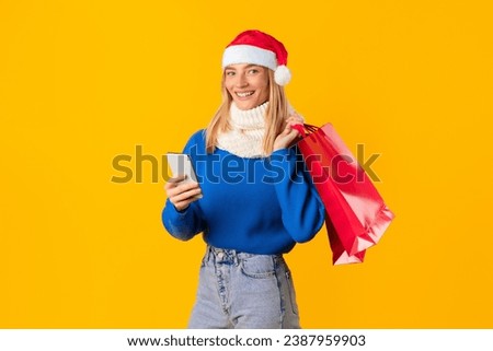 Smiling woman in Santa hat using smartphone while holding red shopping bags, joyfully posing against vibrant yellow background, embodying holiday cheer
