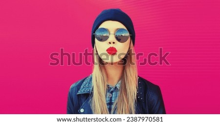 Fashionable portrait of stylish beautiful blonde young woman model blowing her lips with pink lipstick sends sweet kiss in round sunglasses, black rock style leather jacket, hat on city street