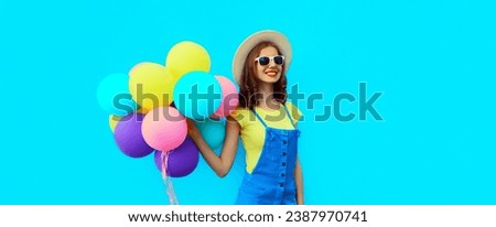 Summer image of happy laughing young woman with bunch of colorful balloons having fun wearing straw hat on blue studio background