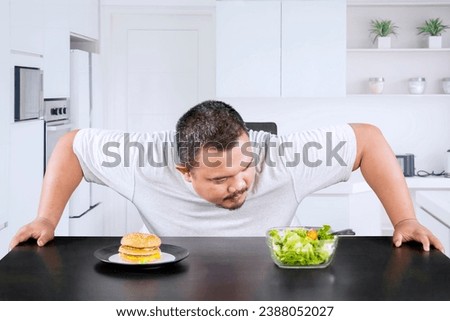 Picture of overweight middle aged man looks confused while choosing a vegetable salad or hamburger in the kitchen