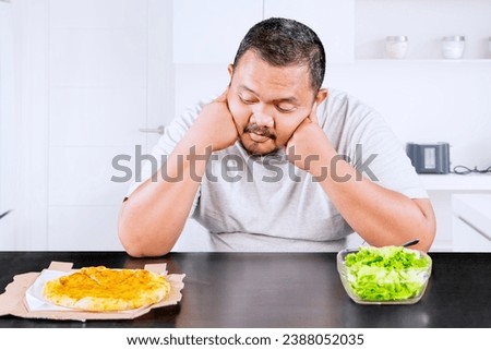 Picture of overweight middle aged man looks confused while choosing a vegetable salad or pizza in the kitchen