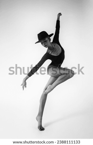 a young ballerina in a black bodysuit shows ballet steps in motion standing on one leg and spreading her hands in a hat