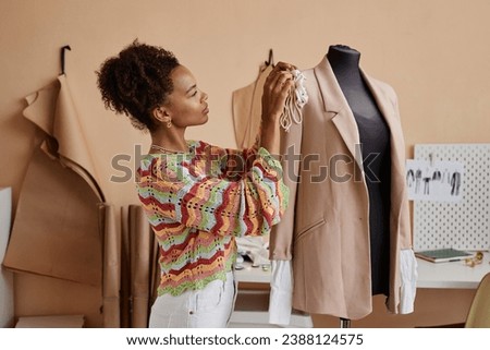 Side view of young female fashion designer sewing handmade decoration on beige blazer hanging on dummy while standing in workshop Royalty-Free Stock Photo #2388124575