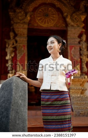 A charming and smiling Thai-Asian woman in a traditional dress with a garland in her hand is visiting a beautiful temple on a Buddhist holy day, looking away from the camera. Thai-culture concept