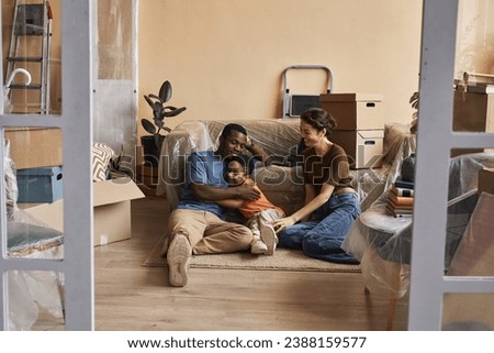 Happy young intercultural family of three relaxing on the floor by couch among boxes with household supplies in spacious living room Royalty-Free Stock Photo #2388159577