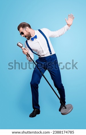 Full size fullbody portrait of famous creative singer in blue pants with suspenders, black glasses, singing hit with open mouth in mic gesture with hand isolated on grey background Royalty-Free Stock Photo #2388174785