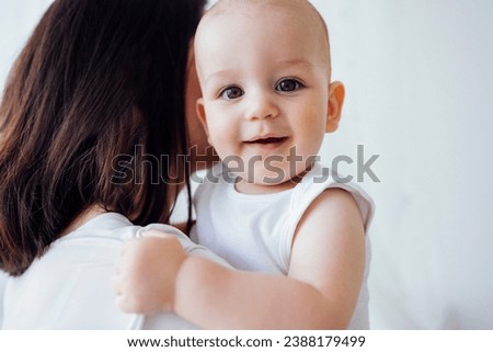 Young beautiful mother hugging her cute baby on cheek. White brunette touches her smiling child with her face. Pretty infant in a white bodysuit with his mom on a light background. Motherhood concept.