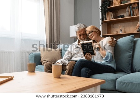 Two elderly people senior couple man and woman looking at a family photo smile and hug while sitting at home on sofa. memories and nostalgia for children went abroad for better life with their family