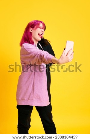 Extraordinary young girl with pink hair and piercing taking selfie with mobile phone against vivid yellow studio background. Concept of youth, self-expression, fashion, emotions, social media