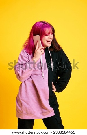 Smiling and laughing young girl with colorful hair and piercing talking on mobile phone against vivid yellow studio background. Concept of youth, self-expression, fashion, emotions, communication