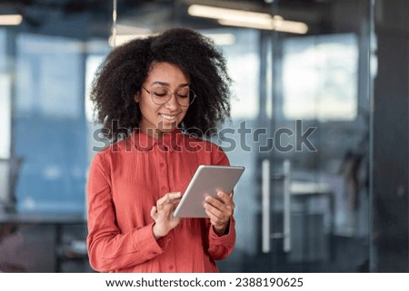 Young successful smiling female programmer inside office at workplace, satisfied woman holding tablet computer in hands, testing new software, business woman with curly hair and shirt.