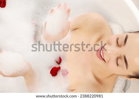Happy woman taking bath in tub with foam and rose petals, top view