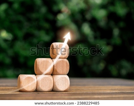 Rising up glowing arrow on empty wooden cube blocks as a business graph or chart steps on table and green leaves background with copy space. Business growth, investment growing and success concepts.