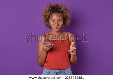 Young attractive ethnic African American woman with mobile phone points finger at you and invites to communicate in instant messengers or social networks stands posing in purple studio. Royalty-Free Stock Photo #2388232851