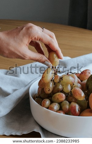 a woman's hand plucks grapes from a white bowl, which stands on a linen kitchen towel on a wooden table