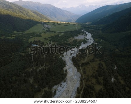 The valley and the town of Mazeri in the distance. Aerial view of the stormy flow of the Dolra River, in the middle of a forest, a hiking trail, a tourist route. Svaneti, Georgia