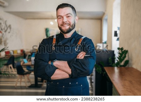 Portrait of happy smiling small business owner dressed in a black chef uniform with an apron in his cozy restaurant hall. Successful people, hard work, consumer cafes and restaurants industry concept. Royalty-Free Stock Photo #2388238265