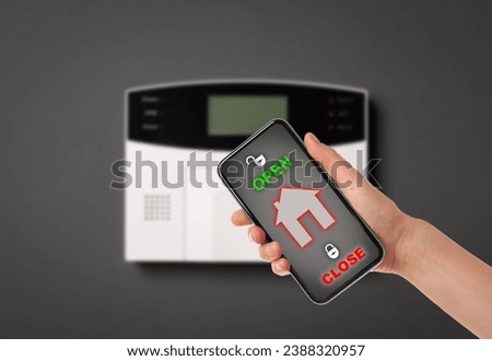Woman operating home alarm system via mobile phone against dark wall with security control panel, closeup. Application interface on device screen