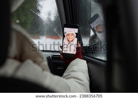Focus on hand of young blond woman holding smartphone with herself on screen while sitting on front seat of car and making selfie