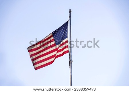 An American flag waving in the wind on a flag pole with a light blue sky background. The national red, white and blue stars and stripes USA flag flying. The US flag is often flown on the 4th of July.
