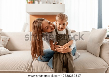 young mother cares and hugs her son on the sofa, woman plays with her child at home, boy 2 years old rejoices and smiles with his parent