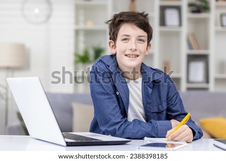 Smiling student boy sitting at a table in a bright room, studying remotely, doing homework, writing in a notebook, smiling, looking at the camera. Royalty-Free Stock Photo #2388398185