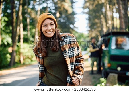 Happy woman spending a weekend camping in the woods and looking at camera. Her friend and travel van are in the background. 
