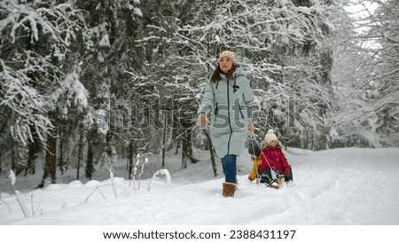 Cheerful playful mom running pulling sled with her kids in winter snowy park.