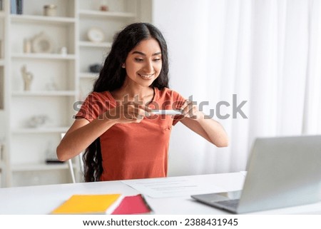 Young indian lady businesswoman using phone, taking photo of documents and sending to client or business partner, sitting at desk with laptop at home office. Modern gadgets and business