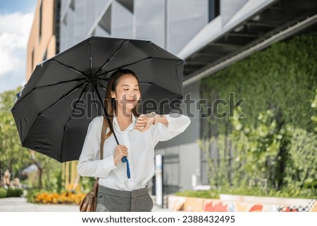 A young Asian businesswoman, shielded by an umbrella, walks to the office under hot sunlight, checking the time during a busy working day. Her commitment to success is evident in her determined look. Royalty-Free Stock Photo #2388432495