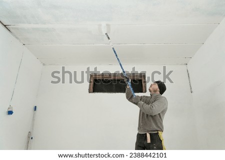 A skilled craftsman meticulously paints a pristine white ceiling using a telescopic roller for precise home improvement. Royalty-Free Stock Photo #2388442101