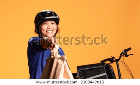 Courier waiting in front of house door for client to pick up takeout meal order, close up. Asian delivery woman offering takeaway food bag to customer, isolated over orange studio background