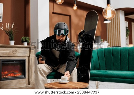 Tourist with cellphone and snowboard equipment in hotel lobby enjoying beverage. Pictured in winter gear, traveler relaxes near fireplace in ski resort lounge area. Ideal for winter holiday themes.