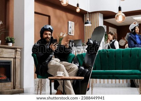 A man seated on sofa browses on his digital phone with snowboard nearby. Caucasian visitor dressed in snow clothing surfing the net using his smartphone. Winter vacation anticipation captured.