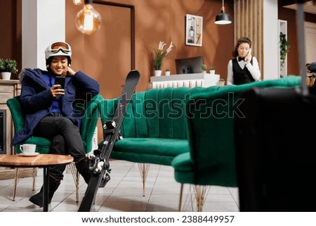Enthusiastic asian lady using her cell phone and waiting in ski hotel lobby for booking procedure for winter vacation. Traveler in wintersports gear with her smartphone in ski resort lounge area.