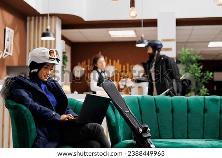 Asian woman wearing winter clothing uses laptop in hotel reception while man is assisted by receptionist. Female guest with personal computer and skiing skis seated on couch in ski mountain resort.