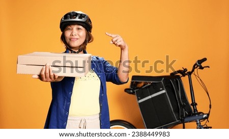 Portrait of joyful professional courier pointing to takeaway pizza boxes. Smiling bike rider showing thumbs up sign while delivering takeout food, isolated over orange studio background
