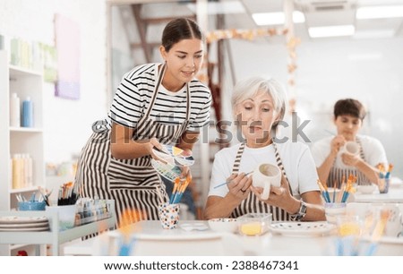 Pleasant young female instructor sharing art of ceramic dishware painting with interested enthusiastic mature woman participating pottery master class Royalty-Free Stock Photo #2388467341
